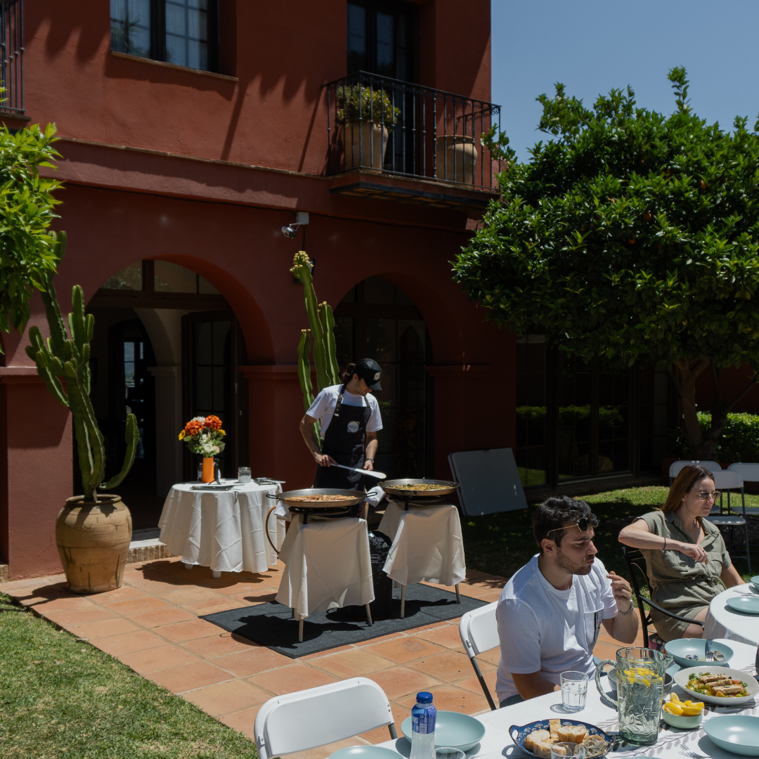 Chef arrocero cocinando al fondo mientras los comensales disfrutan de una mesa preparada para degustar la paella en un evento privado.